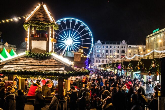 Marché de Noël rempli de monde et illuminé de guirlande et décoration de Noël. Grande roue visible en arrière plan.